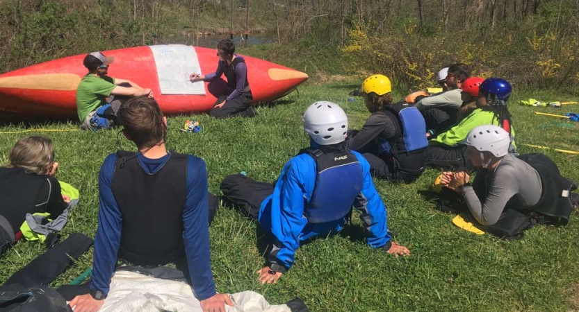 An instructor points to a sheet of paper laid over an upside-down canoe. A group of students rest on the grass, watching.